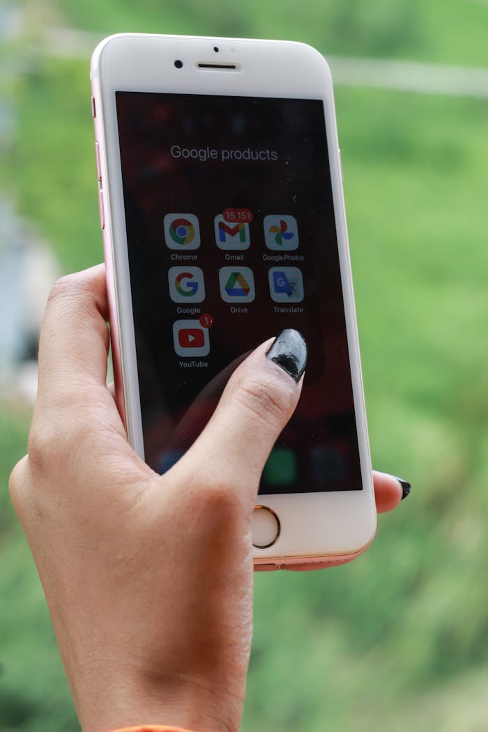 Hand with black nails holding a smartphone showing Google apps outdoors.